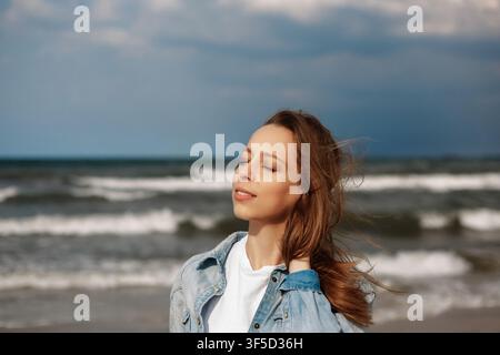 Giovane donna che gode della brezza e del sole sulla spiaggia con gli occhi chiusi, catturando un'atmosfera estiva tranquilla e spensierata, atmosfera pacifica, stile di vita da vacanza estiva spensierata Foto Stock