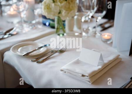 Impostazione elegante del tavolo per un matrimonio o un evento con una cartolina bianca vuota per i nomi, una tovagliolo di lino piegato, posate lucide e un centrotavola floreale morbido per il catering di eventi o la cena di ricevimento Foto Stock