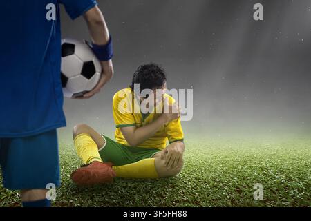 Un giocatore di calcio indonesiano del sud-est asiatico è seduto sul campo erboso dolorante, tenendo la spalla ferita mentre un altro giocatore è vicino e tiene un Foto Stock