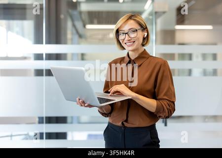Donna d'affari sicura di sé con occhiali e auricolari senza fili, che sorride mentre digita su un laptop, gestisce le attività e rimane connessa in un ambiente aziendale Foto Stock