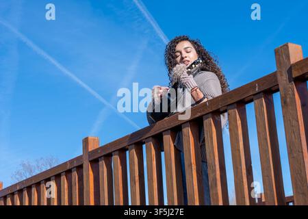 Giovane donna che suona l'ukulele su una ringhiera di legno, godendosi la musica e il cielo azzurro soleggiato Foto Stock