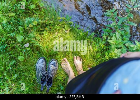 Vista dall'alto di una persona in piedi a piedi nudi su un prato rigoglioso accanto a un fiume che scorre, con scarpe nere posate nelle vicinanze. Momento tranquillo all'aperto in estate, viaggio a Foto Stock