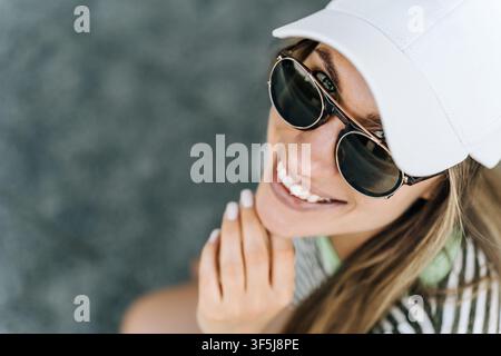Ritratto ravvicinato di una donna sorridente che indossa occhiali da pilota e un berretto bianco, guarda verso l'alto sotto la luce estiva con sicurezza rilassata su uno sfondo morbido Foto Stock