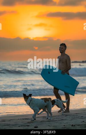 Un surfista porta una tavola blu lungo la spiaggia con due cani al tramonto a Santa Elena, in Ecuador, mentre le onde dell'oceano rotolano sotto un drammatico cielo dorato. Foto Stock