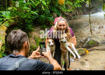 Amici si fermano accanto a un ruscello boschivo su un sentiero delle cascate a Mindo, Pichincha, Ecuador, mentre un viaggiatore fotografa una donna sorridente che tiene due cani. Foto Stock