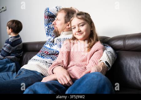 Accogliente scena familiare indoor con un padre e due bambini che si rilassano su un divano in pelle con caldi maglioni di lana, godendosi un momento tranquillo a casa con una luce soffusa della giornata Foto Stock