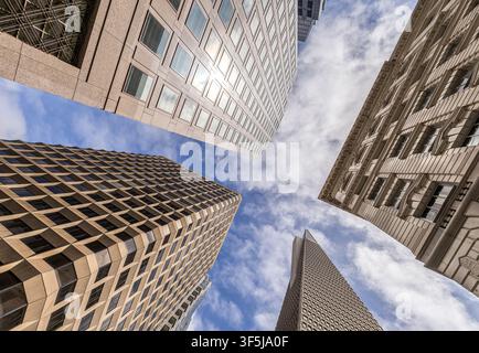 Guarda verso l'alto tra i grattacieli moderni del centro di San Francisco, in California, con facciate geometriche che convergono verso un cielo luminoso e parzialmente nuvoloso. Foto Stock