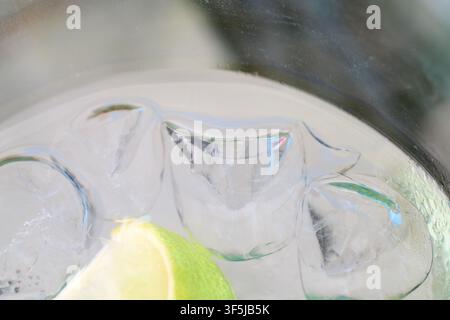 Primo piano di cubetti di ghiaccio cristallini che galleggiano con una vivace fetta di lime all'interno di un bicchiere da cocktail rinfrescante pieno di bevanda gassata. Foto Stock