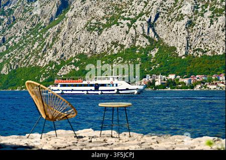 Una vista serena sul mare da un molo di pietra con una sedia e un tavolo in rattan, che guarda verso un grande battello passeggeri sull'acqua blu con montagne sullo sfondo. Foto Stock