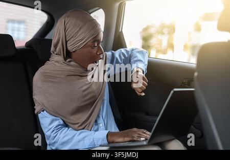 Essere in ritardo. Stressed Businesswoman musulmano nero seduta in taxi e tempo di controllo Foto Stock