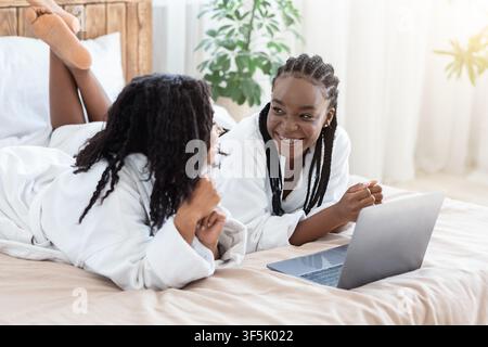 Fidanzate nere positive che si stendano sul letto a casa, usando il laptop Foto Stock
