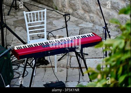 Un primo piano di un tastiere da palco rossa professionale sistemata su un patio in pietra all'aperto. Una sedia bianca elegante si trova dietro lo strumento, circondata da stativi per microfoni e cavi, con foglie verdi che incorniciano lo sfondo. Foto Stock