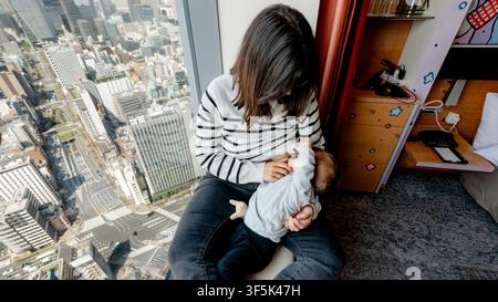 Madre a Minato seduta vicino a una grande finestra che si affaccia sulla skyline di Tokyo, nutre delicatamente suo figlio in una stanza moderna, mostrando cura tenera, stile di vita urbano e f Foto Stock