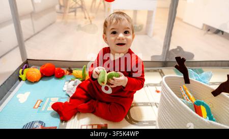 Bambino sorridente in pigiama rosso seduto su un tappeto da gioco colorato, tiene frutti giocattolo dentro un recinto di gioco sicuro a casa, circondato da peluche e un cesto in un br Foto Stock