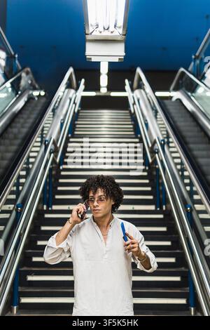 Giovane uomo con occhiali da sole e camicia casuale utilizza uno smartphone mentre è in piedi su una scala tra le scale mobili in una moderna stazione della metropolitana sotto una luce intensa Foto Stock