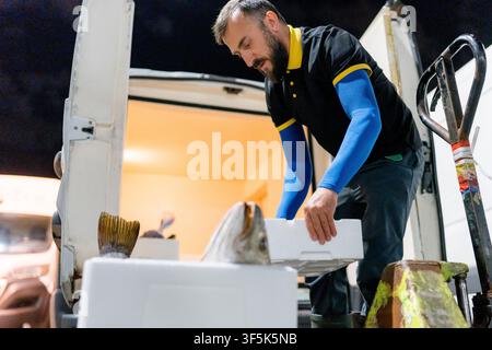 Pescatore scarica pesce fresco in scatole isolate da un furgone di consegna refrigerato di notte, utilizzando un carrello elevatore per il trasporto efficiente di frutti di mare e m Foto Stock