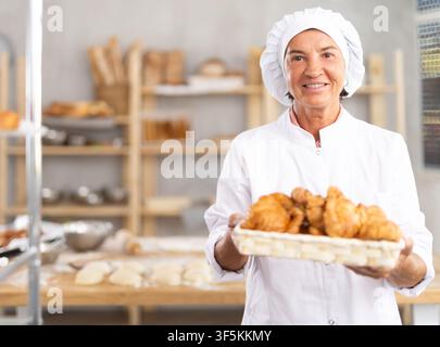 Panettiera anziana sorridente che tiene un vassoio di croissant freschi Foto Stock