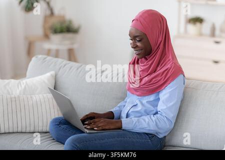 Black Muslim Lady in Hijab lavorando a distanza su laptop AT Casa Foto Stock