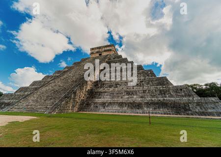 Antica piramide di pietra si erge contro il cielo blu. Nuvole fluttuano sopra la struttura a gradini erosa. Una lussureggiante erba verde copre la base del monumento. Nessuna persona nelle vicinanze Foto Stock