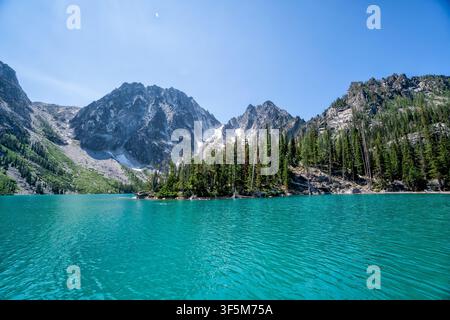 Acqua turchese cristallina rispecchia il cielo blu chiaro. Montagne impervie si ergono maestose dietro la riva boscosa. Il lago calmo brilla sotto il sole luminoso dell'estate. Superficie immobile Foto Stock