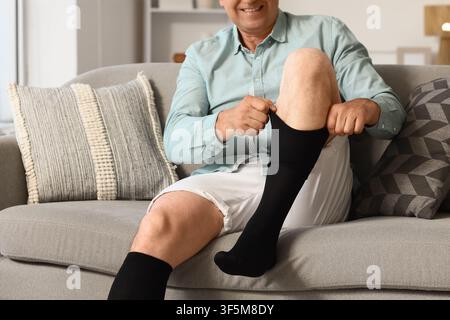 Uomo sorridente che indossa calzini a compressione neri mentre è seduto comodamente su un divano grigio a casa Foto Stock