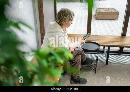 Giovane persona seduta su panchina di legno all'interno di un atrio moderno, controlla lo smartphone accanto a grandi finestre, postura calma, maglione casual, luce naturale del giorno e Foto Stock