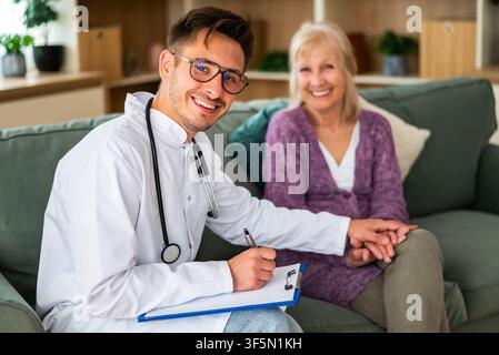 Un operatore sanitario cordiale sorride mentre prende appunti durante una visita a casa, tenendo delicatamente la mano di un paziente anziano per rassicurare e. Foto Stock