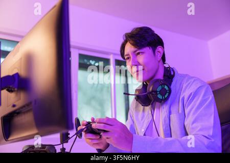 Uomo asiatico che gioca ai videogiochi con un controller a una scrivania illuminata al neon per uno stile di vita da gaming Foto Stock