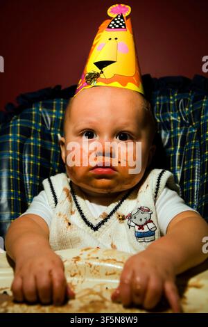 Un bambino di un anno celebra il suo primo compleanno mangiando e indossando una torta gelata Foto Stock