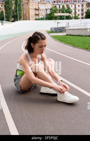 Una bellissima donna atletica è seduta per terra e si allaccia le scarpe prima di un allenamento su una pista. Foto Stock