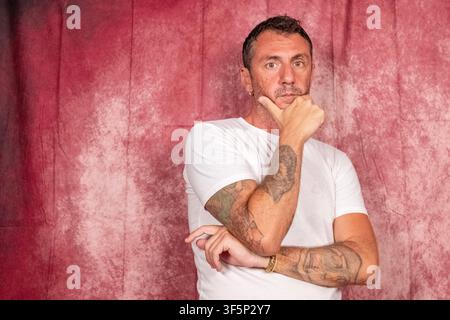 Un uomo rappresentato su uno sfondo rosso, l'uomo ha i capelli corti, una barba corta, indossa una maglietta bianca e ha le braccia tatuate Foto Stock