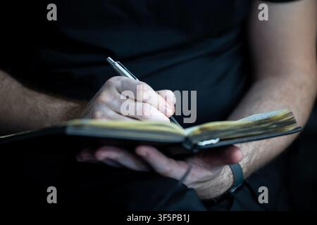 Prima piano di una persona che scrive in un piccolo taccuino con una penna metallica mentre è seduta in un ambiente scarsamente illuminato, indossa una camicia nera con le mani concentrate sul p Foto Stock