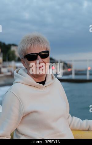 Anziana felice con capelli biondi corti che indossa una felpa casual e occhiali da sole, rilassandosi su un lungomare costiero in un resort europeo. Foto Stock