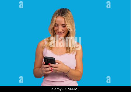 Giovane donna che guarda il telefono, invia un messaggio e sorride su sfondo blu Foto Stock