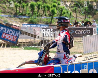 Un pilota di motocross con indumenti protettivi e casco è seduto su una moto da fuoristrada nell'area di partenza di una competizione di corsa fuoristrada. Foto Stock