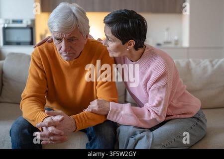 Coppia di anziani seduti sul divano a casa, una donna anziana espressiva consola il marito sopraffatto e sconvolto. Foto Stock