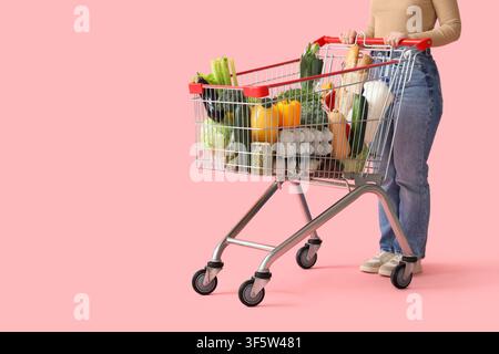 Donna con carrello pieno di diversi prodotti freschi su sfondo rosa Foto Stock