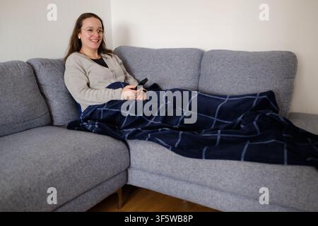 Donna sorridente, accogliente sotto la coperta, tiene un telecomando mentre si rilassa sul divano Foto Stock