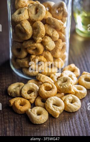 Biscotti taralli italiani dorati e croccanti ammucchiati su un tavolo di legno accanto a un barattolo di vetro, che evocano la pasticceria fatta in casa, momenti rustici di spuntino, momenti accoglienti in cucina e Foto Stock