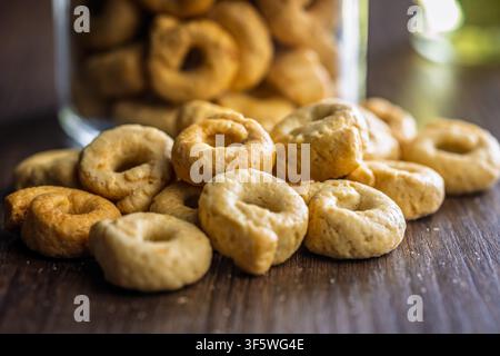 Biscotti taralli italiani dorati e croccanti ammucchiati su un tavolo di legno accanto a un barattolo di vetro, che evocano la pasticceria fatta in casa, momenti rustici di spuntino, momenti accoglienti in cucina e Foto Stock