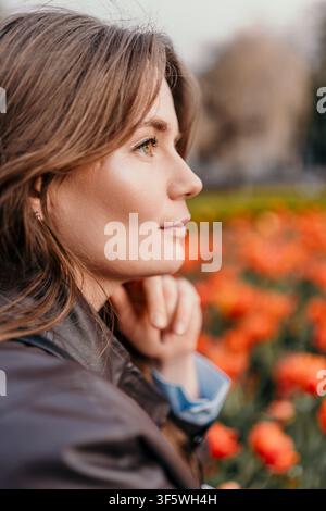 Profilo di donna tulipani Giovane donna con giacca di pelle guarda pensierosa un campo di fiori arancioni in un parco Foto Stock