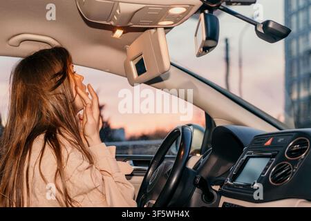 Make-up per auto da donna, giovane donna che controlla la sua pelle nello specchietto parasole all'interno di un veicolo al tramonto Foto Stock