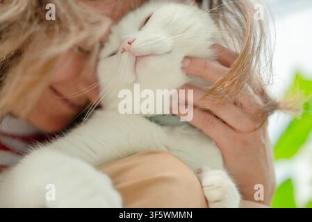 Una donna amorevole tiene stretto il suo gatto bianco, premendo i volti in un abbraccio tenero. Questa proprietaria di animali condivide un momento sereno e affettuoso al chiuso, creando un'atmosfera accogliente di puro conforto e amore. Foto Stock