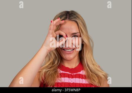 Donna bionda che fa il gesto dell'okay guardando giocosamente attraverso il cerchio formato dalle dita Foto Stock