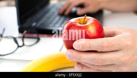 Il concetto di uno spuntino sano al lavoro. Un giovane tiene una mela e lavora su un laptop a casa o al lavoro. Primo piano. Messa a fuoco selettiva. Foto Stock