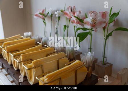 Bancone accogliente del caffè con tovaglioli gialli in portatovaglioli di ottone e fiori di alstroemeria rosa in vasi di vetro su esposizione in legno con luce soffusa. Foto Stock