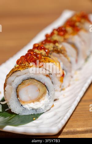 Prima vista di un rotolo di sushi gourmet con tempura di gamberetti fritti, formaggio cremoso, guarnizione grigliata e caviale rosso, servito su piatto bianco con foglia verde su legno Foto Stock