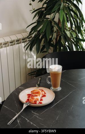 Pancake al formaggio cottage dorato serviti con salsa di fragole e un latte cremoso in un caffè moderno. Colazione o dessert accogliente con un caffè caldo Foto Stock