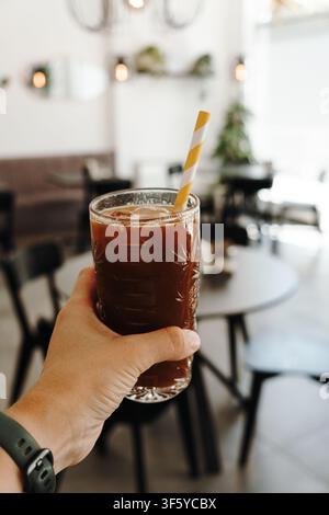 Mano che tiene un bicchiere di caffè freddo con una cannuccia di carta all'interno di un caffè moderno e luminoso. Bevanda fredda rinfrescante con interno sfocato sullo sfondo. Foto Stock