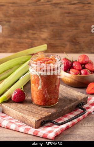 Marmellata fatta in casa di fragole e rabarbaro in barattolo di vetro con ingredienti freschi su un tavolo rustico di cucina Foto Stock